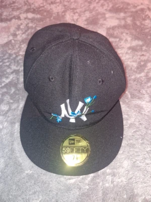 Svart New Era 59FIFTY keps NY - Säljer en svart New Era 59FIFTY keps med klassiskt NY-broderi framtill och blå blommor som detalj. Kepsen har rak skärm och sitter snyggt på huvudet. Perfekt för dig som gillar streetwear och vill sticka ut lite extra.
