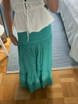 Turkos långkjol med mönster - Säljer en turkos långkjol med fint mönster. Kjolen har en volang nedtill och är lätt och luftig, perfekt för varma dagar. Midjan är elastisk och kjolen har en avslappnad passform. Storlek M men mer som en S