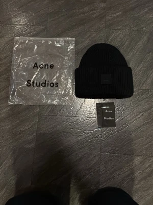 Svart stickad mössa Acne Studios - Svart ribbstickad mössa från Acne Studios med uppvikt kant och den klassiska ansiktsloggan framtill. Mössan är tillverkad i ett mjukt stickat material som håller dig varm och har en stilren, minimalistisk look. Perfekt accessoar för kyliga dagar.