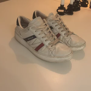 Moncler vita sneakers med röd och blå detalj - Säljer ett par snygga vita sneakers från Moncler med klassiska röd och marinblå ränder på sidan och logga på hälen. Skorna har rund tå, snörning och är tillverkade i skinn. Perfekta för dig som gillar stilrena och sportiga skor med exklusiv känsla.