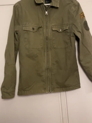 Grön overshirt från Zara  - Snygg militärgrön. Jackan har två stora bröstfickor med knappar och stängs med dragkedja framtill. Tillverkad i bomull, perfekt för lager på lager och chill stil. Tror inte denna säljs längre. Pris går o diskutera 
