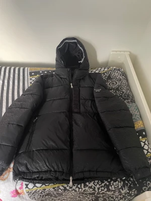 Svart pufferjacka Alessandro Zavetti - Säljer en svart pufferjacka från Alessandro Zavetti med huva och diskret logomönster över hela jackan. Jackan har dragkedja framtill, två sidofickor med dragkedja och en snygg badge på ärmen. Perfekt för kalla dagar när du vill hålla stilen.