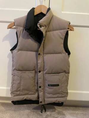 Beige dunväst från Canada Goose 2XS - Säljer en beige vintage dunväst från Canada Goose i storlek 2XS. Västen har två stora fickor framtill, hög krage och stängs med både dragkedja och knappar. Utsidan är i polyester och bomull, fodrad med nylon och fylld med ankdun. Prutningsbar