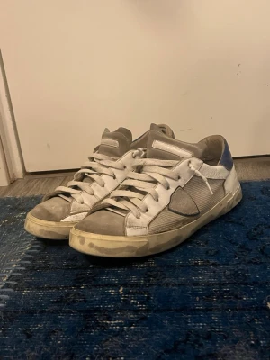 Philippe Model sneakers i vitt och blått - Säljer ett par Philippe Model sneakers med vit och grå ovandel i skinn och mesh, blå detaljer på hälen och klassisk snörning. Snygg rund tå och platt sula, perfekt för dig som gillar stilrena och sportiga skor med fransk vibe. 