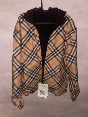Burberry rutig vindjacka med huva - Säljer en ikonisk Burberry vindjacka med klassiskt beige, svart och rött rutmönster. Jackan är vändbar med svart insida, har huva och dragkedja framtill. Tillverkad i lätt polyester, perfekt för dig som vill ha en clean och tidlös look.