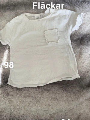 Vit t-shirt med volangficka från ZARA - En basic vit t-shirt från ZARA med korta ärmar och en söt liten ficka med volangdetalj på bröstet. Perfekt till jeans eller shorts för en enkel och clean look.🌸 Fläckar. Storlek 98