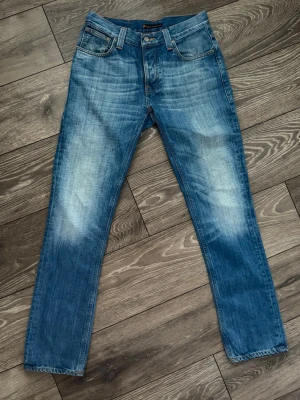 Blå jeans från Nudie Jeans - Säljer ett par klassiska blå jeans från Nudie Jeans med snygg slitning och kontrastsömmar. Modellen är grim Tim och har raka ben, fem fickor och ikonisk broderad detalj på bakfickorna. Tillverkade i slitstarkt bomullstyg och har normal passform.