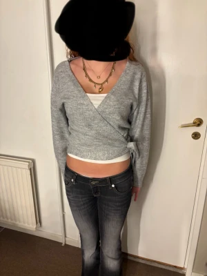 Grå omlott kofta med knyt - Säljer en ljusgrå omlott kofta med knyt i sidan. Koftan har v-ringad hals och långa ärmar, och är stickad i mjukt material. Perfekt att styla med ett linne under och jeans för en chill look.