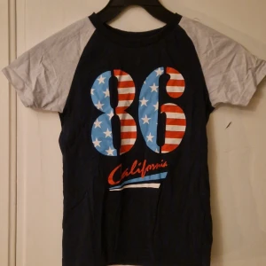 Mörkblå t-shirt med tryck från Name It - Cool mörkblå t-shirt från Name It med kontrasterande ljusgrå ärmar. Framsidan har stort tryck med siffran 86 i amerikanskt flaggmönster och texten 'California' i rött. Perfekt för en avslappnad och sportig look. Hund finns i hemmet. Kolla in mina andra annonser för samfrakt! 