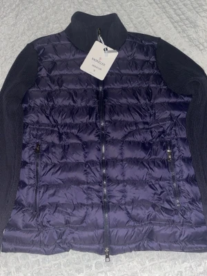 Mörkblå dunväst från Moncler - Säljer en mörkblå dunväst från Moncler med quiltad front och två dragkedjeförsedda fickor. Västen har stickade partier på sidor och krage för extra komfort och stil. Klassisk Moncler-logga på insidan och coolt serietryck. Perfekt för lager-på-lager.