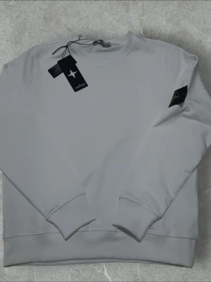 Vit sweatshirt från Stone Island - Vit långärmad sweatshirt från Stone Island med klassisk rund hals och ribbade muddar. Tröjan har den ikoniska Stone Island-loggan på vänster ärm och är tillverkad i mjuk bomull. Perfekt för dig som gillar stilrena och exklusiva plagg.