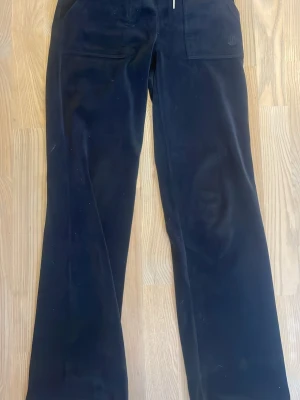 Juicy couture set svart - Säljer ett par svarta mjukisbyxor och en zip up tröja från Juicy Couture i mjuk sammet. Byxorna har dragsko i midjan, raka ben och stora fickor med lock och silverknappar. Perfekta för chill dagar och riktigt skön passform.