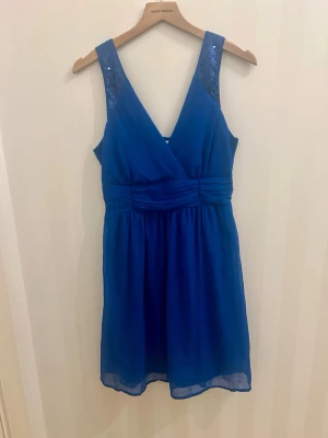 Blå omlottklänning från Vero Moda - Supersnygg blå omlottklänning från Vero Moda breda axelband dekorerade med paljetter. Klänningen har markerad midja och lätt utställd kjol i luftig polyester. Perfekt för dig som vill ha något festligt! 