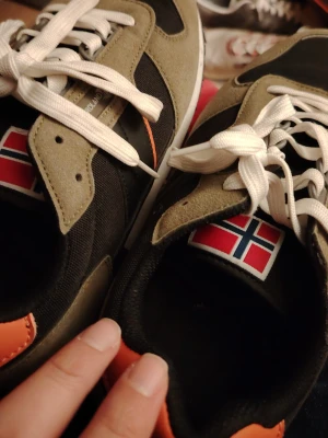 Nordland sneakers med norsk flagga - Säljer ett par snygga sneakers från Nordland med norsk flagga på plösen. Skorna är svarta med olivgröna detaljer i mocka, vita skosnören och en orange detalj vid hälen. De har en klassisk retrostil med platt sula och svart/vit yttersula.