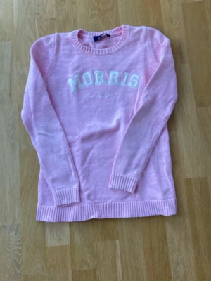Rosa stickad tröja från Morris - Säljer en ljusrosa stickad tröja från Morris med vit text på bröstet. Tröjan har rund hals, ribbade muddar och nederkant samt långa ärmar. 