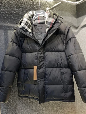 Svart pufferjacka med huva - Säljer en svart pufferjacka med huva och dragkedja. Jackan har rutigt foder i grått, svart och rött, samt två fickor med dragkedja framtill. Perfekt för kalla dagar och riktigt skön att bära. Stängs med både dragkedja och knappar.