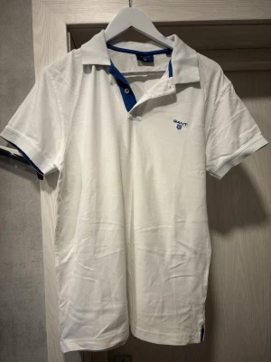 Vit pikétröja från Gant - Klassisk vit pikétröja från Gant med blå detaljer vid kragen och knäppningen. Tröjan har korta ärmar och Gant-logga broderad på bröstet. Tillverkad i mjuk bomull som känns skön mot huden. Perfekt till jeans eller shorts för en clean look.