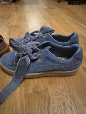 Blå Puma Suede sneakers med rosett - Säljer ett par blå Puma Suede sneakers i storlek 39. Skorna har breda sammetsband som snörning och en snygg rosett framtill. Ovandelen är i mjuk mocka och sulan är platt och beige. Guldiga detaljer vid snörningen och Puma-logga på sidan.
