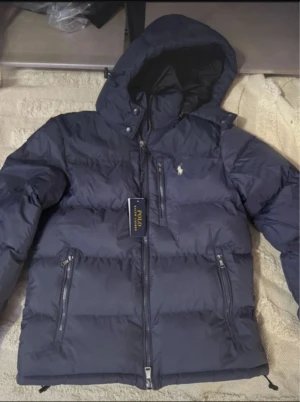 Mörkblå dunjacka från Polo Ralph Lauren - Snygg mörkblå dunjacka från Polo Ralph Lauren med huva och dragkedjor på fickorna. Jackan har ett klassiskt puffer-utseende och Polo-loggan broderad på bröstet. Perfekt för kalla dagar och har en clean, sportig vibe.