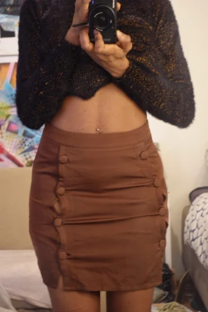 Brun kortkjol med knappar - Säljer en snygg brun kortkjol med tight passform. Kjolen har dekorativa knappar längs sidorna och en diskret slits nedtill. Materialet känns mjukt och lätt, perfekt för dig som gillar stilrena detaljer. Dragkedja i sidan för enkel på- och avtagning.