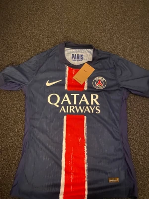 PSG matchtröja D.Doué #14 Nike - Paris Saint-Germain fotbollströja med D.Doué och nummer 14 på ryggen. Tröjan är marinblå med en bred röd och vit rand på framsidan, PSG-emblem på bröstet och Champions League-märke på ärmen. Tillverkad i lätt, ventilerande Dri-FIT-material.