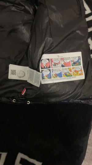 Svart dunväst från Moncler - Svart, glansig dunväst från Moncler med huva och två dragkedjeförsedda fickor. Västen har quiltad design och Moncler-logga på bröstet. Materialet är polyester och dun, perfekt för kyliga dagar. Snygg och stilren look med klassisk Moncler-detalj. Med nfc-scan