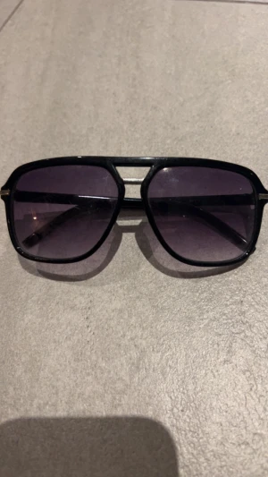 Svarta pilotbågar solglasögon - Snygga solglasögon med svarta pilotbågar och mörka tonade glas. Ramen är i plast med metall-detaljer vid sidorna och en klassisk dubbelbrygga. Glasen har en lätt övergång från mörkt till ljusare ton. Perfekt för dig som gillar retrostil och vill sticka ut.