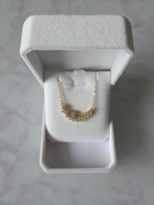 Halsband med glittriga stenar - Supersnyggt halsband i guld med en böjd hängande detalj dekorerad med små glittriga stenar. Kedjan är tunn och elegant, och hängsmycket har flera rader med gnistrande stenar som ger extra bling. Perfekt för dig som gillar att sticka ut med accessoarer.