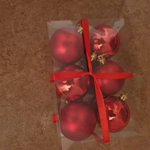 Julkulor - Denna bild visar en förpackning med sex röda julgranskulor, både blanka och matta, med guldfästen. Kulorna är snyggt förpackade i en transparent ask med röd rosett. Perfekt för att dekorera granen inför julen. Inga synliga defekter eller slitage.