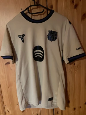 FC Barcelona – Lamine Yamal #10 – Special Edition - En exklusiv FC Barcelona-tröja i beige/guld med Lamine Yamal #10 på ryggen. Tröjan är sprillans ny, aldrig ens testad. Detaljer: * Officiell Barça-design * Lamine Yamal namn + nummer * UNHCR-logga * Specialmärken på ärmen (bl.a. 2025-märke) * Storlek: M Perfekt för samlare eller Barça-fans som vill ha något unikt.   Kan skickas eller hämtas.                                            (Shorts följer med)