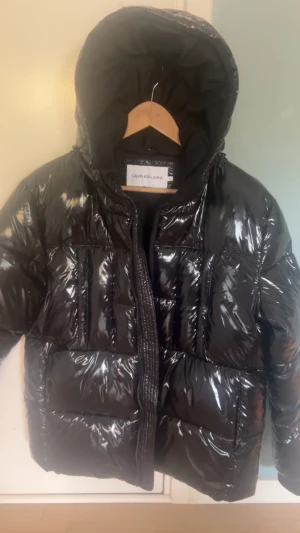 M. Svart glansig pufferjacka Calvin Klein Nyskkck  - Storlek M. Svart pufferjacka från Calvin Klein Jeans med glansig finish och stor huva. Jackan har dragkedja framtill och breda, vadderade paneler som ger en riktigt fet look. Perfekt för kalla dagar när du vill sticka ut lite extra. Nyskick. 