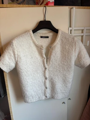 Vit kortärmad topp från Gina Tricot - Superfin vit topp från Gina Tricot i fluffigt, stickat material. Modellen är croppad med korta ärmar och rund halsringning. Knäpps framtill med silvriga knappar som ger en söt touch. Perfekt att styla med jeans eller kjol för en trendig look.