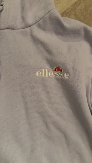 Ljuslila hoodie från Ellesse - Snygg ljuslila hoodie från Ellesse med broderad logga på bröstet. Tröjan har huva med snörning, stor magficka och ribbade muddar. Perfekt för chill dagar och streetstyle. Mjuk bomullskvalitet som känns skön mot huden.