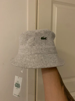 Grå bucket hat från Lacoste - Bucket hat i ljusgrått från Lacoste, med klassisk krokodillogga broderad framtill. Tillverkad i mjuk frotté som ger en chill vibe och skön känsla. Perfekt för att lyfta din streetstyle och hålla dig cool under soliga dagar.