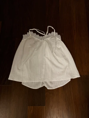 Vit linnetopp med spetsdetaljer - Säljer en söt vit linnetopp med tunna axelband och spetsdetaljer längs v-ringningen. Toppen är lätt och luftig med en flowy passform, perfekt för varma dagar. Materialet är mjukt och har en diskret struktur som ger extra känsla. Spetsen ger en romantisk touch.