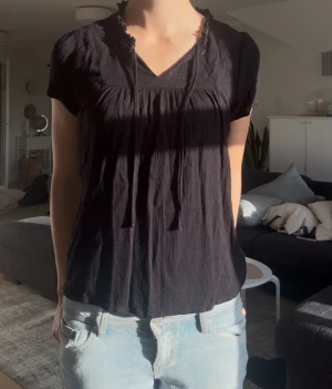 Svart blus med knyt och volang - Svart blus med korta puffärmar och v-ringning med knytband. Halsen har en söt volangkant och tyget är lätt skrynklat för en chill vibe. Blusen är luftig och har en avslappnad passform, perfekt till jeans eller kjol.