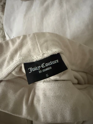 Vita mjukisbyxor från Juicy Couture - Supermjuka vita mjukisbyxor från Juicy Couture i velour med bred resår i midjan och snygg ficka bak med JC-detaljer på knapparna. Perfekta för en chill och trendig look.