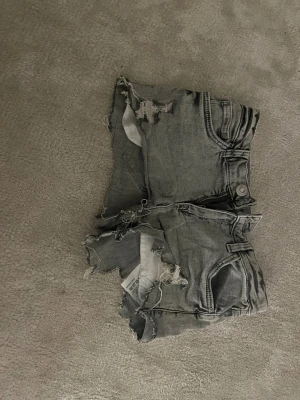 Grå denimshorts med råa kanter - Säljer ett par gråa denimshorts från Kappahl i storlek 146. De har en cool, sliten look med råa och fransiga kanter samt slitningar framtill. Klassisk femficksmodell med knapp och dragkedja. Perfekta för dig som gillar streetstyle och edgy vibe. Egenklippta drf säljs billigare men väldigt fina annars