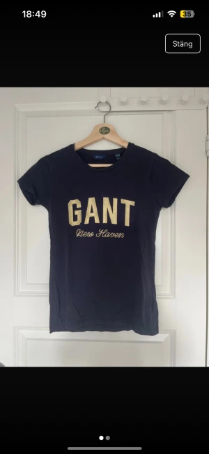 Mörkblå GANT t-shirt med guldtryck - Snygg mörkblå t-shirt från GANT med stort guldigt tryck på bröstet där det står 'GANT New Haven'. Klassisk rund halsringning och korta ärmar. Tillverkad i mjuk bomull som är skön att ha på sig.