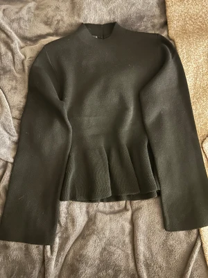 Svart peplumtröja med knappar bak - Svart peplumtröja från H&M med lång ärm och hög hals. Tröjan har snygga knappar längs ryggen och ett ribbat peplum-parti nedtill som ger en cool siluett. Perfekt för dig som gillar stilrena och moderna plagg.