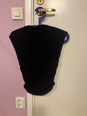 Svart ärmlös topp med drapering - Snygg svart topp med draperad effekt på sidorna och ärmlös design. Toppens form är lite loose och den har en rund halsringning. Perfekt för dig som gillar stilrena och enkla plagg med en twist. Materialet känns mjukt och följsamt mot kroppen.