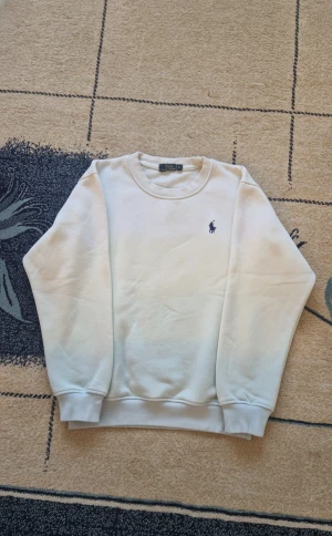 Vit sweatshirt från Polo Ralph Lauren - Snygg vit sweatshirt från Polo Ralph Lauren med klassisk rund hals och det ikoniska lilla blå Polo-logot på bröstet. Mjuk och skön bomullsmix, ribbade muddar vid ärmar och nederkant. Perfekt för chill dagar och enkel att matcha med jeans eller joggers.