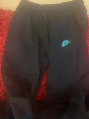 Svarta mjukisbyxor från Nike S - Svarta mjukisbyxor från Nike i storlek S med turkos Nike-logga på benet. Byxorna har snörning i midjan och dragkedja på fickan. Perfekta för chill eller träning, mjukt material och relaxed fit. Säljer nu eftersom jag inte använder mjukisbyxor. Byxorna är helt nya och aldrig använts