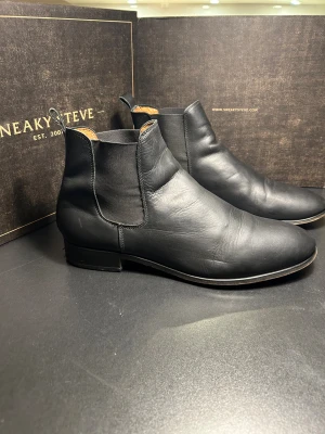 Svarta chelsea boots Shoe the bear - Snygga svarta chelsea boots från Shoe the bear i mjukt skinn med elastiska paneler på sidorna och rund tå. Klassisk siluett med låg klack och dragflik bak för enkel påtagning. Perfekta för dig som gillar stilrena och tidlösa boots.
