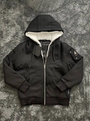  Moose Knuckles Bunny - Svart jacka/hoodie från Moose Knuckles med vit fluffig fuskpäls på insidan av huvan. Jackan har dragkedja framtill, snörning vid huvan och en snygg metall-logga på ärmen. Ribbad mudd vid ärmslut och nederkant, samt praktiska fickor framtill.