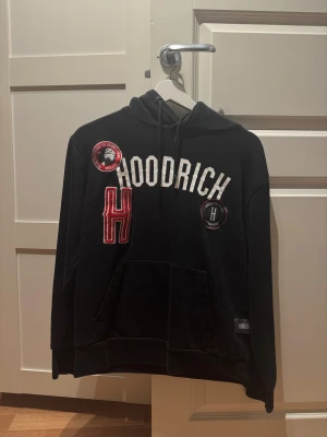 Svart hoodie från Hoodrich med tryck - Svart hoodie från Hoodrich i storlek S med stor vit logga och röda detaljer på bröstet. Tröjan har huva med snörning, känguruficka och flera broderade patches. Perfekt för dig som gillar streetwear och vill sticka ut med coola detaljer.