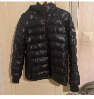 Svart pufferjacka från Peak Performance - Säljer en svart pufferjacka från Peak Performance med huva och ribbade muddar. Jackan har en glansig yta, horisontella quiltade sektioner och dragkedja framtill. Perfekt för kalla dagar och har en modern, sportig vibe.