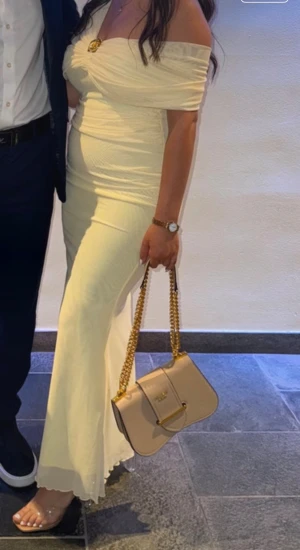 Ljusgul långklänning med blomdetalj - Supersnygg ljusgul långklänning med omlott-design och offshoulder. Klänningen har en unik guldfärgad blomma som brosch framtill och är figurnära med lätt volangkant nertill. Materialet är mjukt och tunt. Perfekt för dig som vill sticka ut med en elegant look. Endast använd en gång 