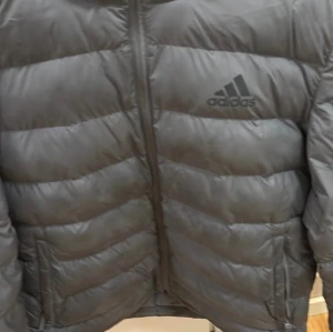 Grå pufferjacka från Adidas - Säljer en svart pufferjacka från Adidas med huva och dragkedja framtill. Jackan har ett stilrent, quiltat mönster och Adidas-loggan på bröstet. Perfekt för kalla dagar och har en modern, sportig vibe. Passar dig som gillar streetwear och vill hålla dig varm.