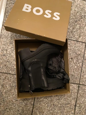 Svarta boots från BOSS i skinn - Säljer ett par svarta boots från BOSS i äkta skinn med chunky platt sula och rund tå. Skorna har elastiska paneler på sidorna och snygga detaljer i guld vid hälen. Perfekta för dig som gillar stilrena och robusta skor med modern känsla.